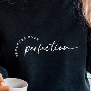 May include: Black sweatshirt with white text that reads "Progress over perfection" in a cursive font.