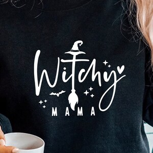 May include: Black sweatshirt with a white graphic design that says "Witchy Mama" with a witch's hat, broom, and stars.