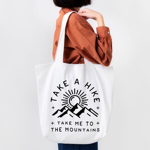 May include: White tote bag with black text and graphic. The text reads "Take a Hike + Take Me To The Mountains". The graphic is a sun with mountains behind it.