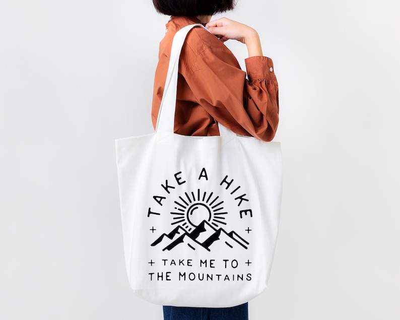 May include: White tote bag with black text and graphic. The text reads "Take a Hike + Take Me To The Mountains". The graphic is a sun with mountains behind it.