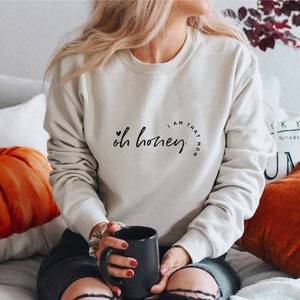 Puede incluir: Sudadera beige con la frase "Oh honey, I am that mom" en escritura negra. La sudadera es de cuello redondo. Una taza negra es sostenida en las manos de la persona que lleva la sudadera.