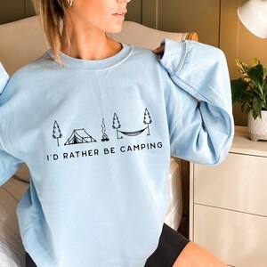 May include: A light blue sweatshirt with a black graphic of a tent, campfire, hammock, and trees. The text "I'd rather be camping" is printed below the graphic.