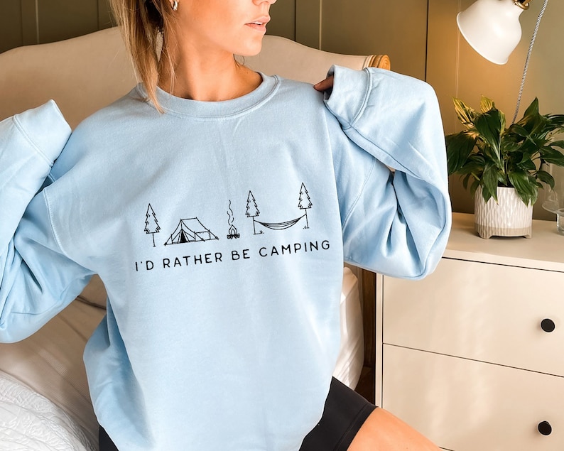 May include: A light blue sweatshirt with a black graphic of a tent, campfire, hammock, and trees. The text "I'd rather be camping" is printed below the graphic.