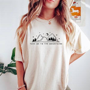 May include: A white t-shirt with a black graphic of mountains, trees, a crescent moon, and stars. The text "Take me to the mountains" is printed below the graphic.