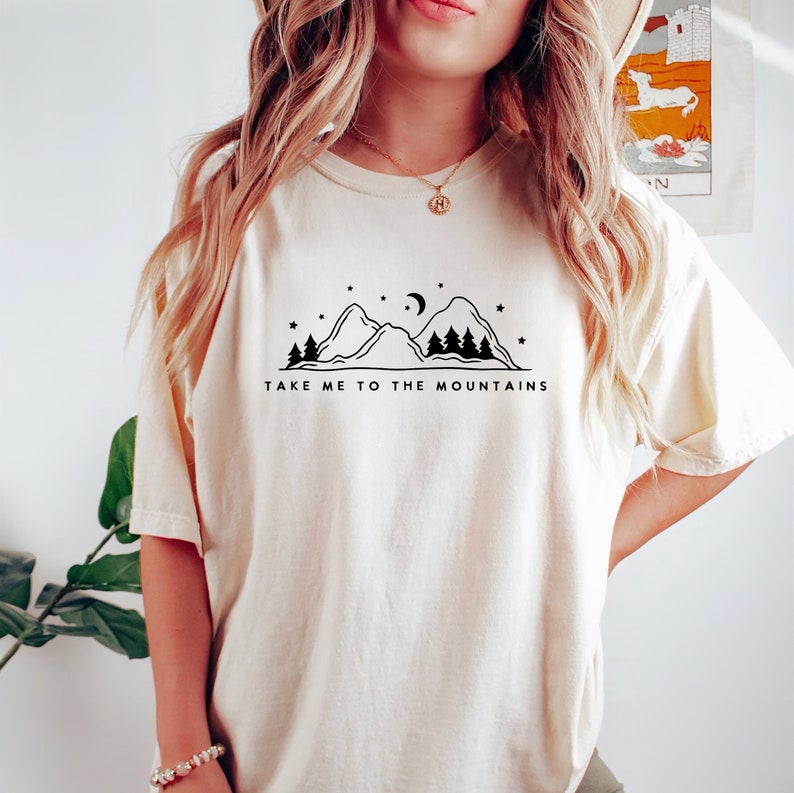 May include: A white t-shirt with a black graphic of mountains, trees, a crescent moon, and stars. The text "Take me to the mountains" is printed below the graphic.