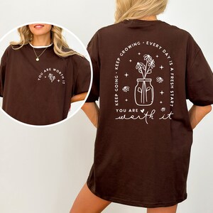 May include: Brown oversized t-shirt with a white graphic print on the back that says "Keep going. Keep growing. Every day is a fresh start. You are worth it." The graphic features a jar with flowers and stars.