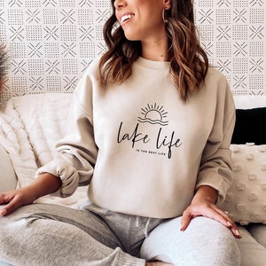 Peut inclure: Un sweat-shirt beige avec un graphique noir d'un soleil couchant sur l'eau et le texte "lake life is the best life".