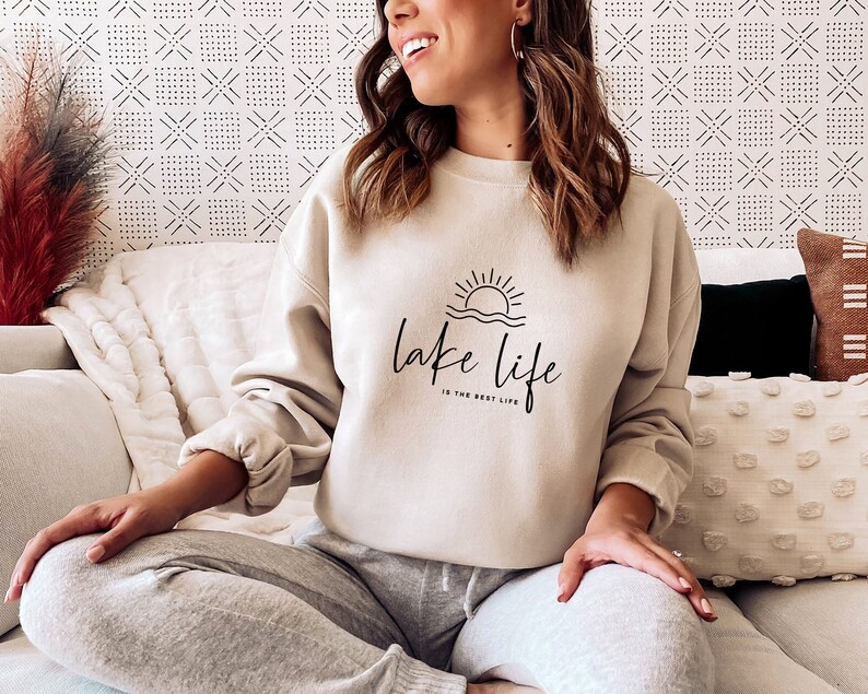 Peut inclure: Un sweat-shirt beige avec un graphique noir d'un soleil couchant sur l'eau et le texte "lake life is the best life".