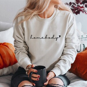 Op de afbeelding: Een wit sweatshirt met de tekst "homebody" en een klein hartje in zwarte letters.