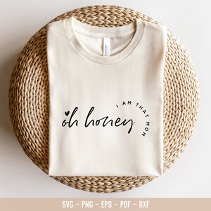 Puede incluir: Camiseta color crema con el texto "oh honey" y "I am that mom" en escritura negra. La camiseta est&aacute; doblada sobre una superficie de mimbre tejido. El dise&ntilde;o incluye un peque&ntilde;o s&iacute;mbolo de coraz&oacute;n.