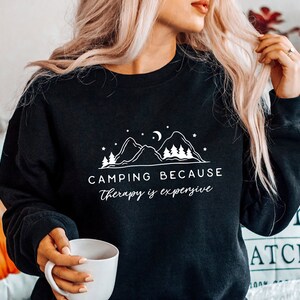 May include: Black sweatshirt with a white graphic design of mountains, a crescent moon, and stars. The text reads "Camping because therapy is expensive."