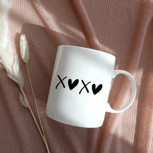 May include: White ceramic mug with black lettering that says "XOXO" with hearts between the "X"s. The mug is sitting on a pink fuzzy surface.
