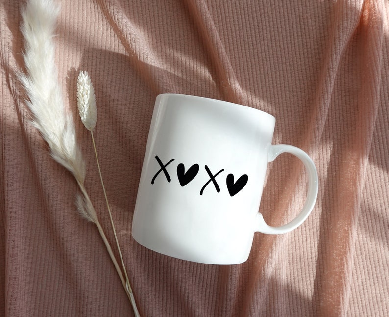 May include: White ceramic mug with black lettering that says "XOXO" with hearts between the "X"s. The mug is sitting on a pink fuzzy surface.