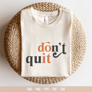 May include: A white t-shirt with the text "don't quit" in black and orange lettering. The shirt is folded and displayed on a woven mat.