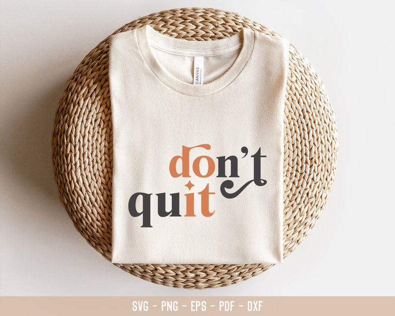 May include: A white t-shirt with the text "don't quit" in black and orange lettering. The shirt is folded and displayed on a woven mat.