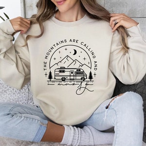 May include: A cream-colored sweatshirt with a black graphic design of a camper van driving through a mountain landscape with the text "The Mountains Are Calling And I Must Go".