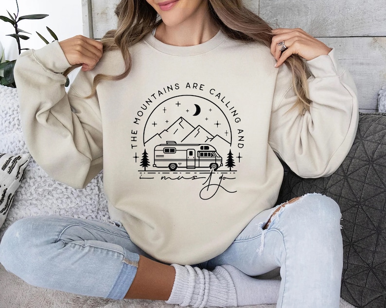 May include: A cream-colored sweatshirt with a black graphic design of a camper van driving through a mountain landscape with the text "The Mountains Are Calling And I Must Go".
