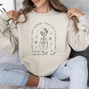 May include: A beige sweatshirt with black text that reads "Keep going - Keep growing. Every day is a fresh start. You are worth it." The text is in a circle around a black and white illustration of a jar with flowers.