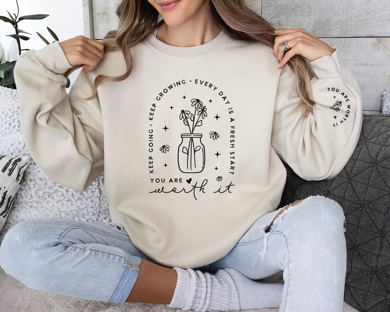 May include: A beige sweatshirt with black text that reads "Keep going - Keep growing. Every day is a fresh start. You are worth it." The text is in a circle around a black and white illustration of a jar with flowers.
