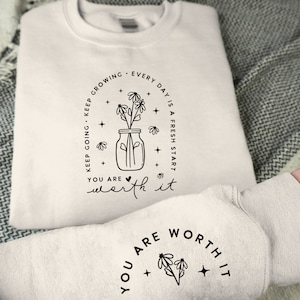 May include: White sweatshirt with a black graphic design of a mason jar with flowers and the text "KEEP GROWING. KEEP GOING. EVERY DAY IS A FRESH START. YOU ARE WORTH IT." The same text is repeated on the sleeve of the sweatshirt, but without the mason jar graphic.