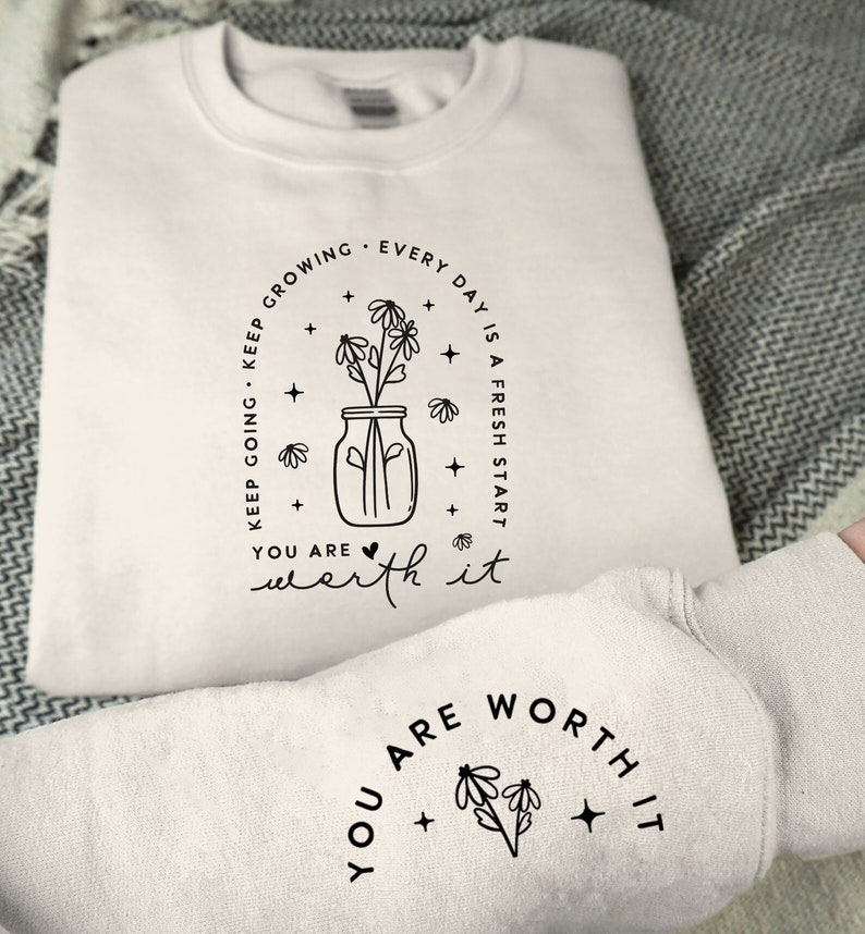 May include: White sweatshirt with a black graphic design of a mason jar with flowers and the text "KEEP GROWING. KEEP GOING. EVERY DAY IS A FRESH START. YOU ARE WORTH IT." The same text is repeated on the sleeve of the sweatshirt, but without the mason jar graphic.