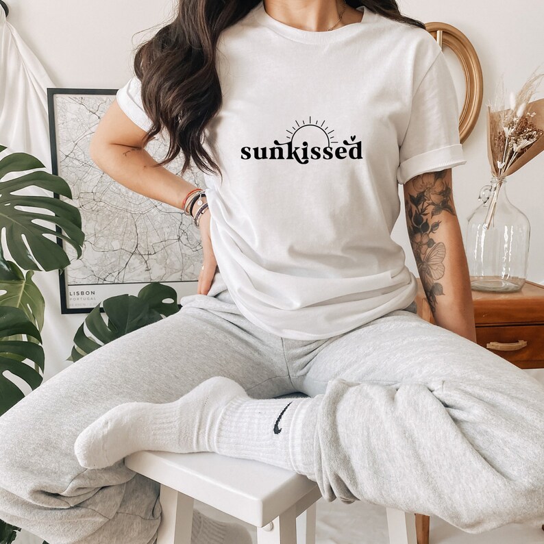 May include: A white t-shirt with a black graphic of a sun and the word "sunkissed". The model is wearing grey sweatpants and white socks.