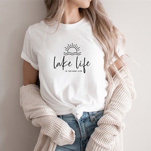 Peut inclure: T-shirt blanc avec un graphique noir d'un soleil couchant sur l'eau et le texte "lake life is the best life".