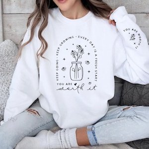 May include: White sweatshirt with a black graphic design featuring a jar of flowers and the text "Keep growing. Every day is a fresh start. You are worth it."