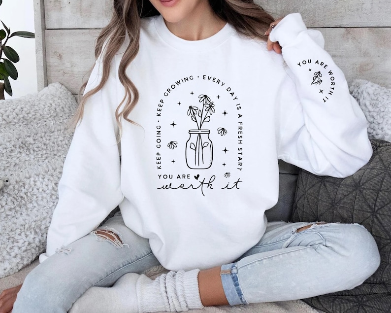 May include: White sweatshirt with a black graphic design featuring a jar of flowers and the text "Keep growing. Every day is a fresh start. You are worth it."
