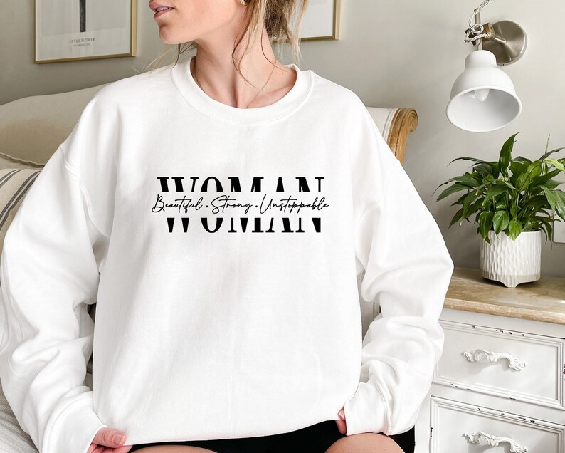 May include: White sweatshirt with black text that reads "WOMAN Beautiful. Strong. Unstoppable WOMAN".