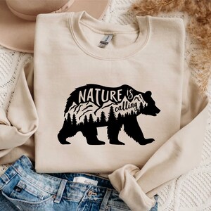 May include: A beige sweatshirt with a black bear silhouette design. The bear has mountains and trees inside its outline. The text "NATURE is calling" is printed above the bear.