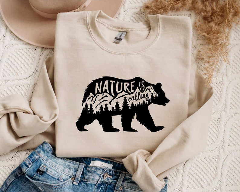 May include: A beige sweatshirt with a black bear silhouette design. The bear has mountains and trees inside its outline. The text "NATURE is calling" is printed above the bear.