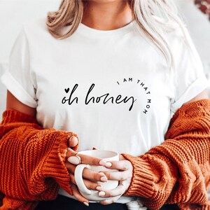 Puede incluir: Camiseta blanca con la frase "Oh honey, I am that mom" en escritura negra. La camiseta se combina con un su&eacute;ter de punto trenzado color &oacute;xido. Una taza blanca se sostiene en las manos.