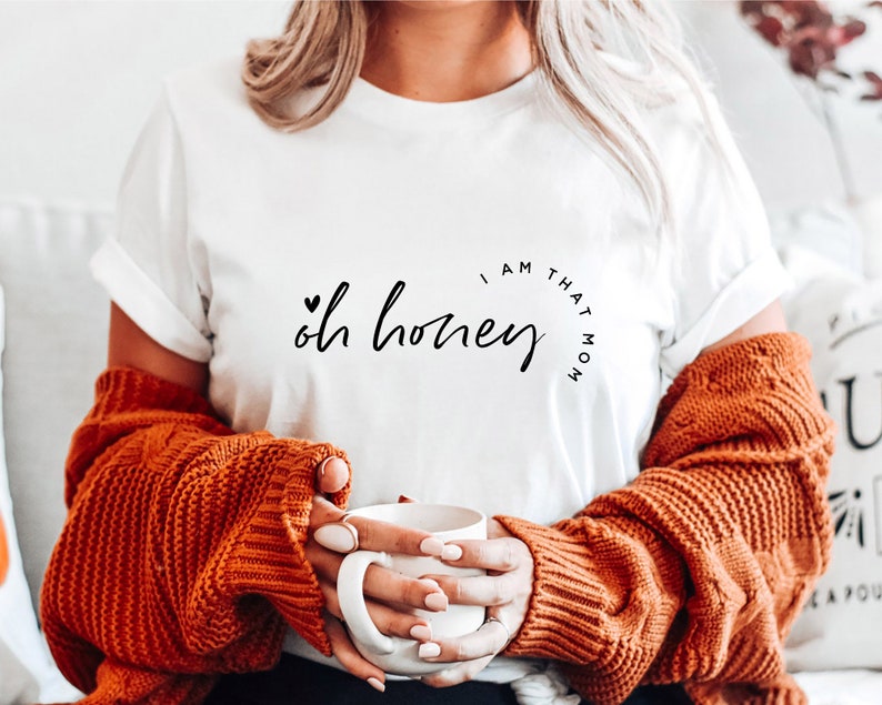 Puede incluir: Camiseta blanca con la frase "Oh honey, I am that mom" en escritura negra. La camiseta se combina con un su&eacute;ter de punto trenzado color &oacute;xido. Una taza blanca se sostiene en las manos.