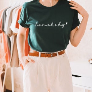 Op de afbeelding: Een donkergroen T-shirt met de tekst "homebody" in witte cursieve letters en een klein wit hartje.
