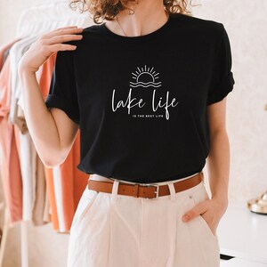 Peut inclure: T-shirt noir avec un graphique blanc d'un soleil couchant sur l'eau et le texte "lake life is the best life".
