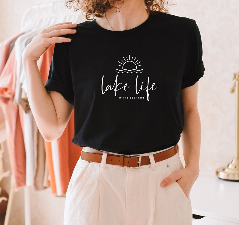 Peut inclure: T-shirt noir avec un graphique blanc d'un soleil couchant sur l'eau et le texte "lake life is the best life".