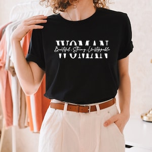 May include: Black t-shirt with white text that reads "WOMAN Beautiful. Strong. Unstoppable. WOMAN"