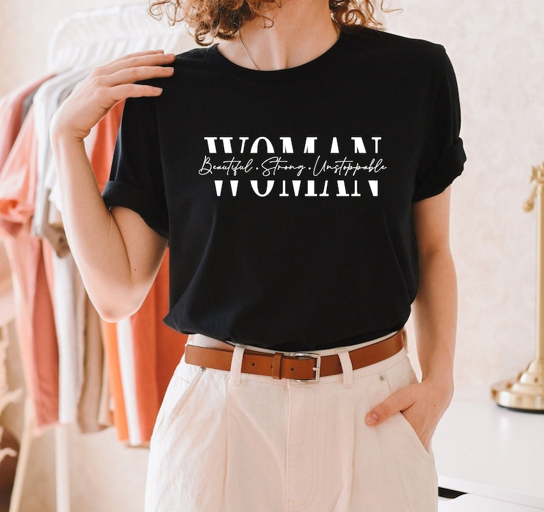 May include: Black t-shirt with white text that reads "WOMAN Beautiful. Strong. Unstoppable. WOMAN"