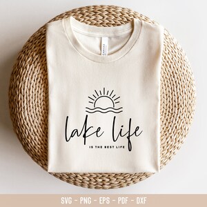 Peut inclure: Un t-shirt blanc avec un motif graphique noir repr&eacute;sentant un soleil couchant sur un lac avec le texte "lake life is the best life".