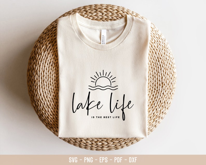 Peut inclure: Un t-shirt blanc avec un motif graphique noir repr&eacute;sentant un soleil couchant sur un lac avec le texte "lake life is the best life".