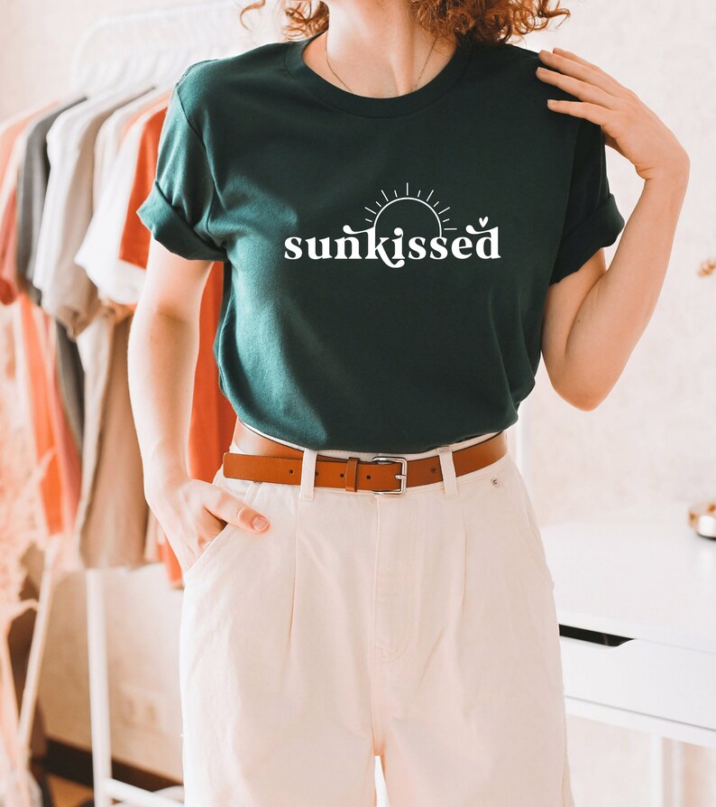 May include: A dark green t-shirt with a white graphic of a sun and the word "sunkissed". The model is wearing the shirt with white pants and a brown belt.