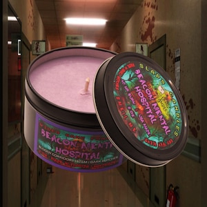 May include: A purple candle in a black tin with a label that reads "Video Game Fans Beacon Mental Hospital Bloody Corridors | Stem | Dark Butcher 8 oz. Made in Florida Handmade"