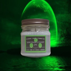 Op de afbeelding: Een grijze glazen kaars pot met een bruin deksel. Het etiket luidt "VIDEO GAME FANATIC SAM FISHER'S COVERT OPS DARK PLUMILIGHT AND STEALTHY MUSK INFILTRATED OFFICE 5 OZ I MADE IN FLORIDA I HANDMADE".