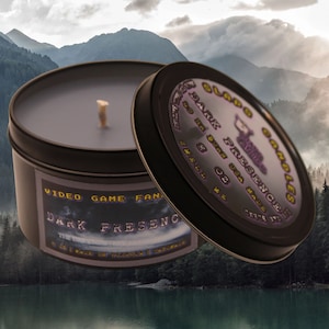 Op de afbeelding: Een zwarte kaarsblik met een paars en gouden etiket met de tekst "GLAD'S CANDLES DARK PRESENCE" en "VIDEO GAME FAN", tegen een achtergrond van een mistig bergmeer.