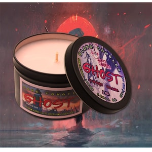 May include: A black candle tin with a pink candle inside. The tin has a red, white, and black graphic with the text "SLAPS CANDLES GHOST 8 oz. SMELL ME" and "GAME FANATIC GHOST ROSEMARY | FRESH | BLOOD | SAGE HANDMADE IN FLORIDA".