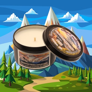 Może przedstawiać: Czarne i srebrne pudełko na świecę z białą świecą w środku. Etykieta głosi "Obado Gadgets Laps Candles Strange Bay Town Soy Wax Blend 8 oz Smell Me". Pudełko stoi na zielonym i brązowym tle z górami i drzewami.
