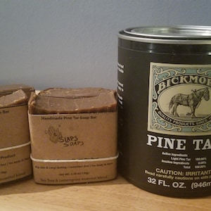Può includere: Tre barre di sapone al catrame di pino marrone fatte a mano con un'etichetta di carta marrone che recita "Handmade Pine Tar Soap Bar" e "SLAPS SOAPS". Il sapone è accanto a una lattina nera di Bickmore Pine Tar, che è un prodotto per il trattamento del legno. La lattina ha un'etichetta con un'illustrazione in bianco e nero di un cavallo che tira un carro.
