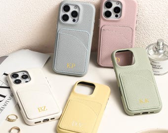 May include: Five leather phone cases in various colors: light gray, pink, white, yellow, and green. Each case has a cardholder pocket and gold-colored initials. The cases are arranged on a white surface with a magazine and perfume bottle in the background.