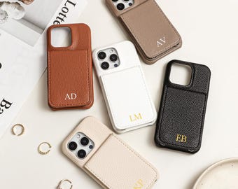May include: A collection of personalized phone cases in various colors, including brown, white, beige, and black. Each case features a unique initial in gold lettering. The cases are arranged on a white surface with a magazine and rings.